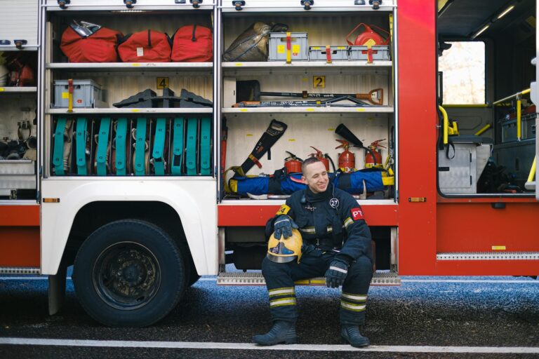 Firefighter in uniform sitting by fire truck, surrounded by equipment, smiling.